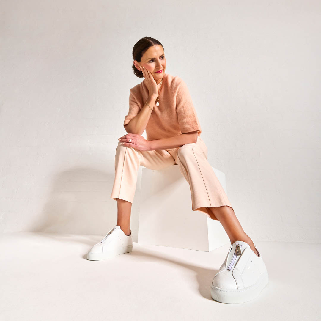 Woman sitting on a white stool wearing a pink outfit with white estella sneakers against a plain background