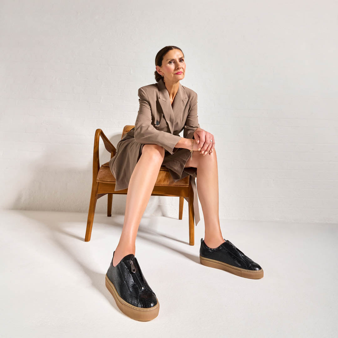 Woman sitting on a wooden chair wearing black Estella shoes with brown soles against a white background