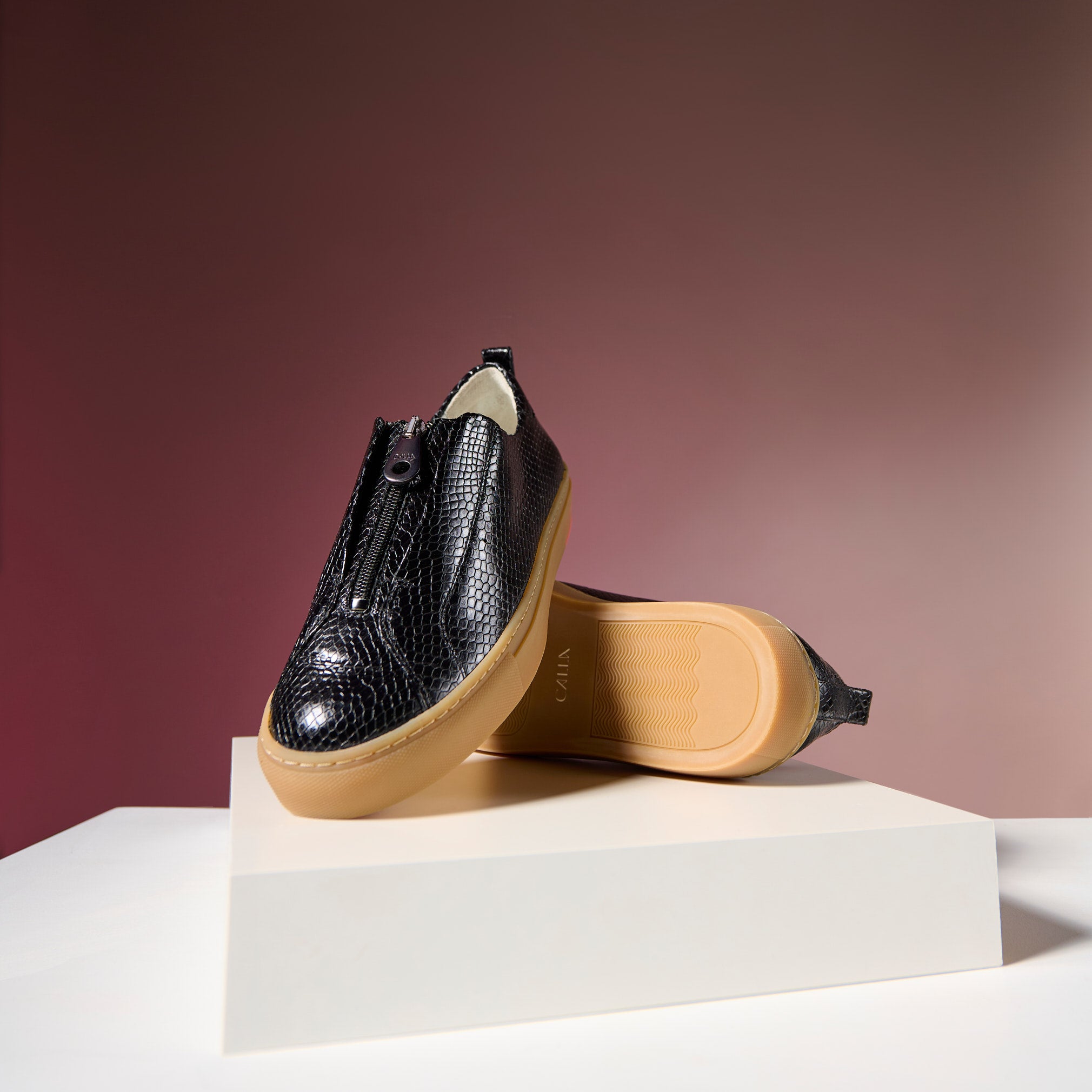 Black textured Estella sneaker with tan sole on a white platform against a red background