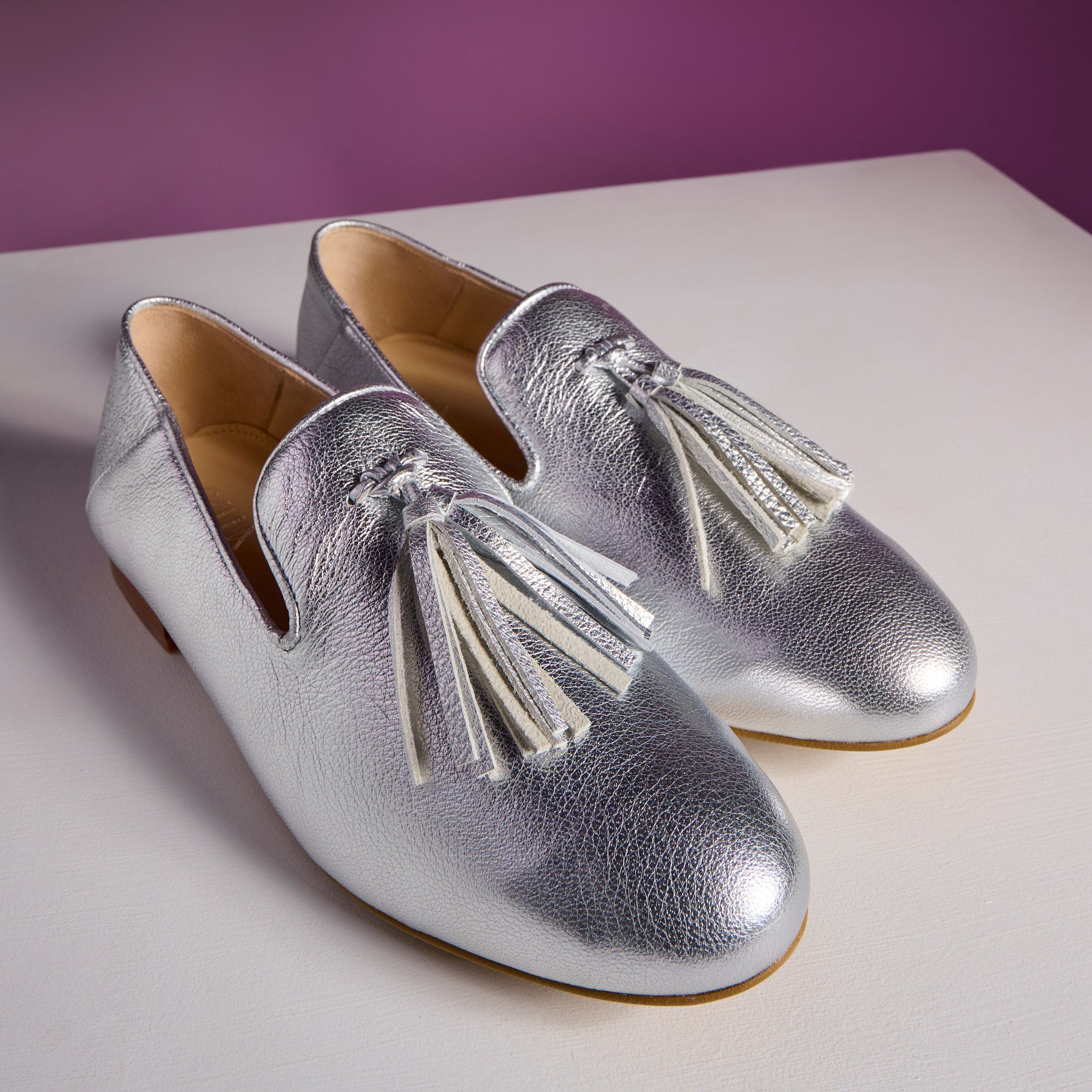 Silver loafers with tassels on a white surface with a purple background