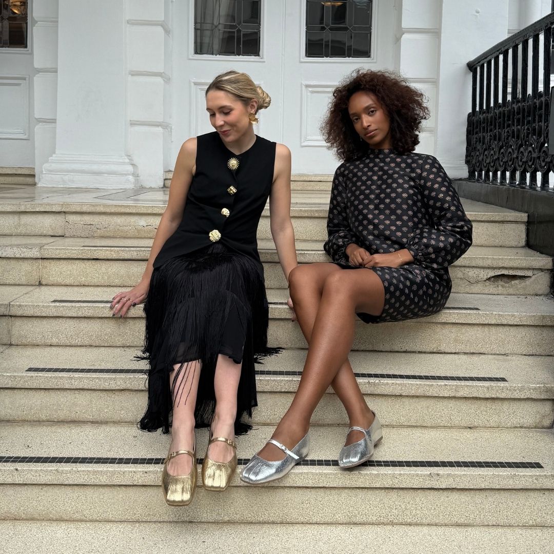 Two women sitting on steps wearing black dresses with gold buttons and silver shoes.