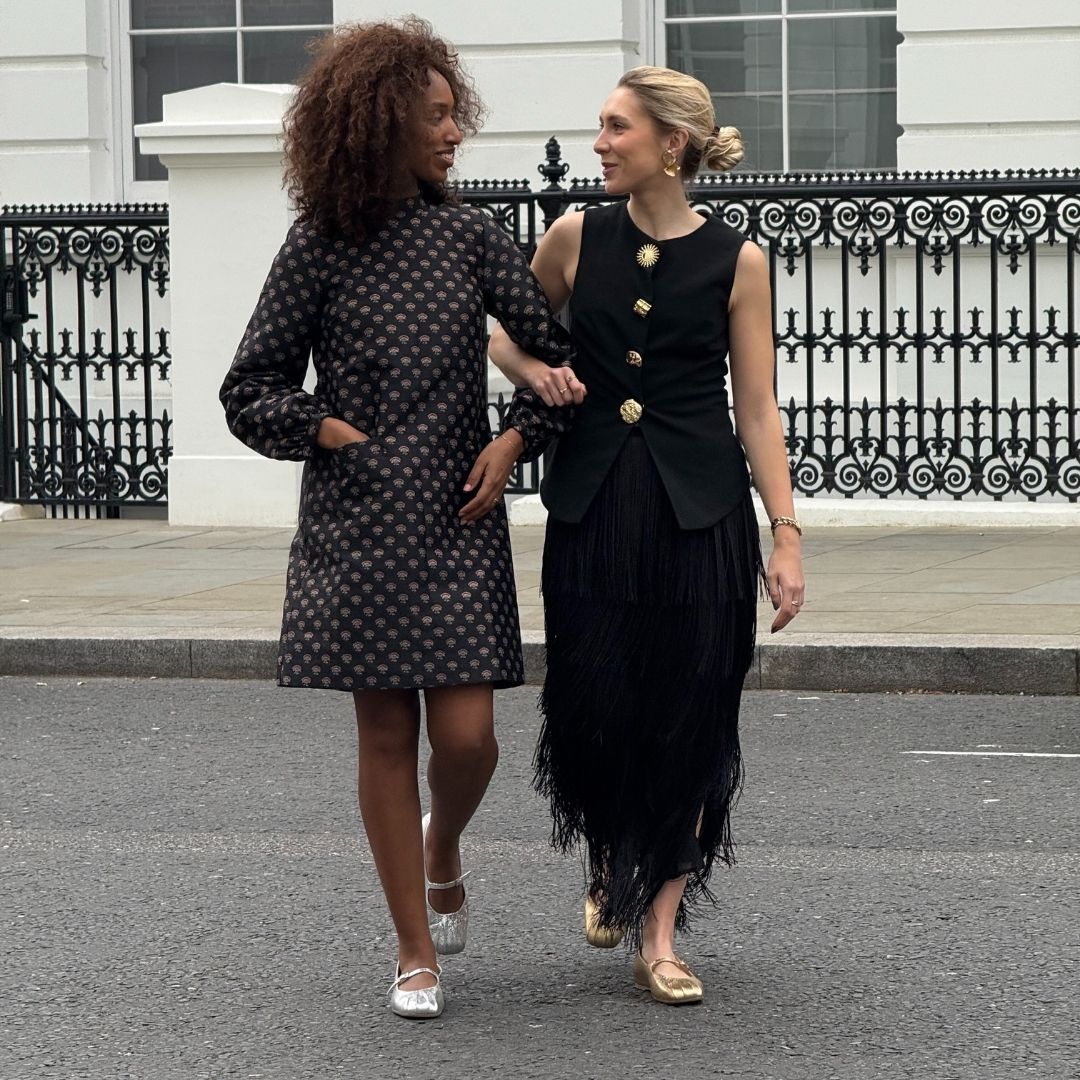 Two women walking together on a street, one in a patterned dress and the other in a black dress with gold buttons.