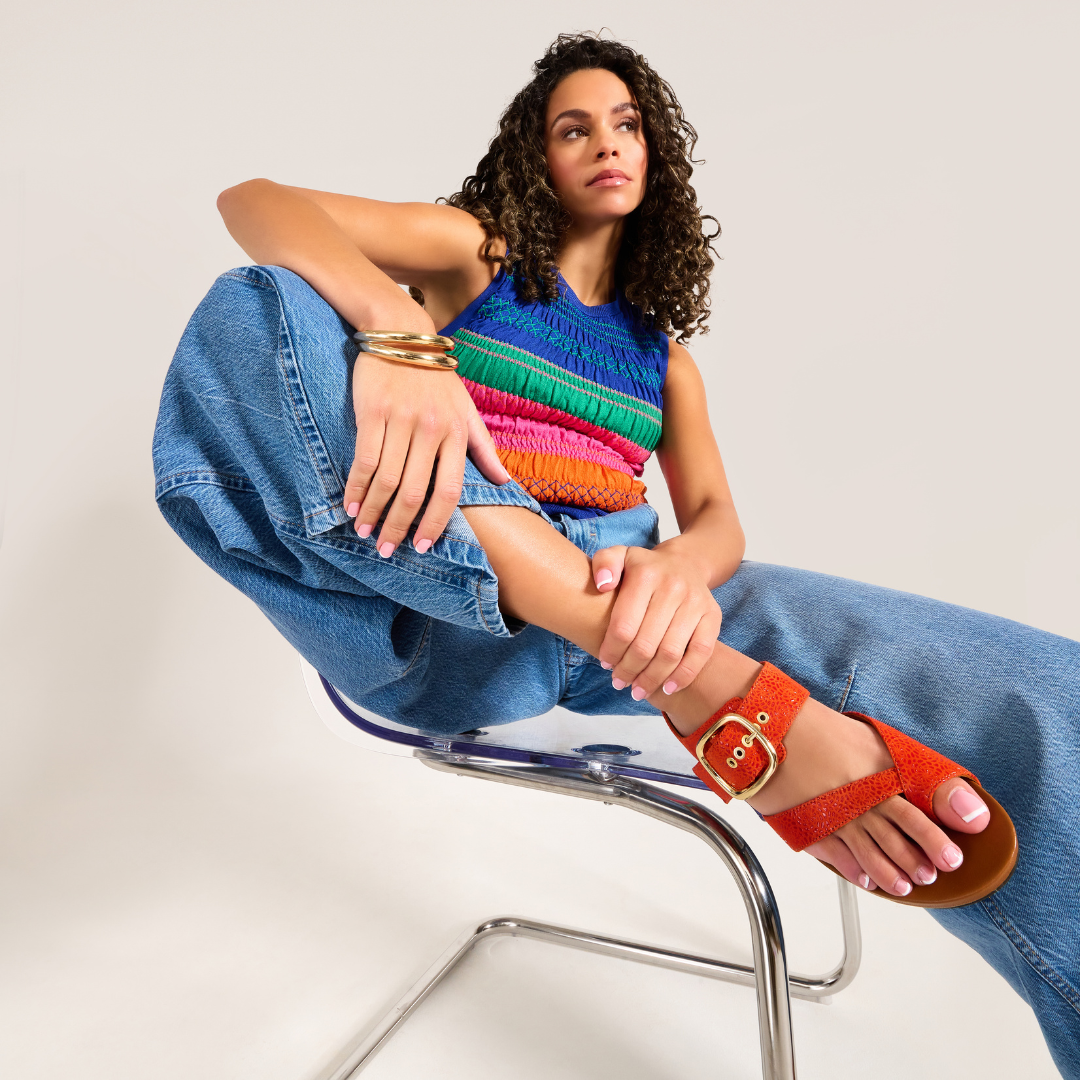 Woman sitting on a chair wearing colorful sandals, denim jeans, and a multicolored top.