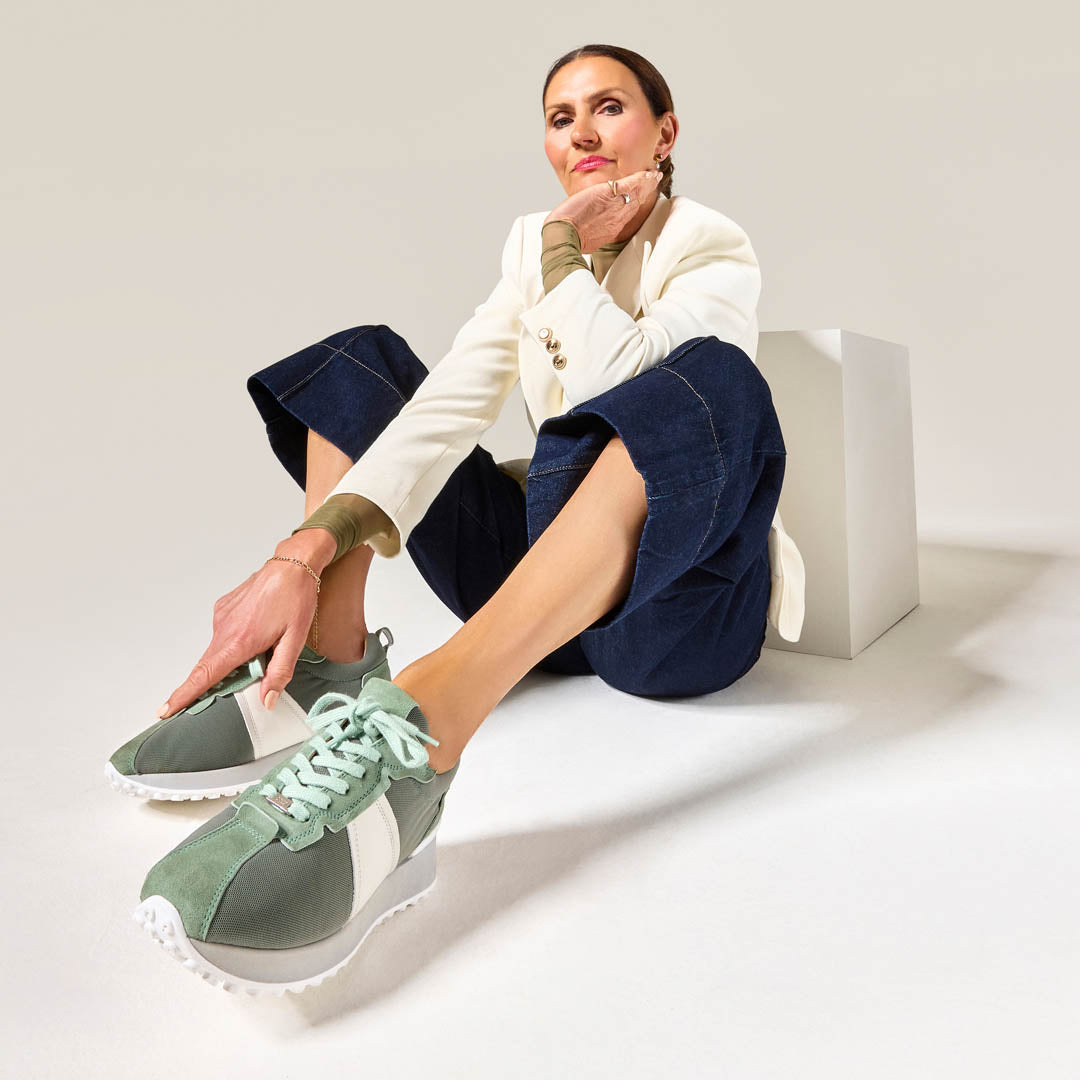 Woman sitting on a white block wearing green cosmos sneakers with a neutral background
