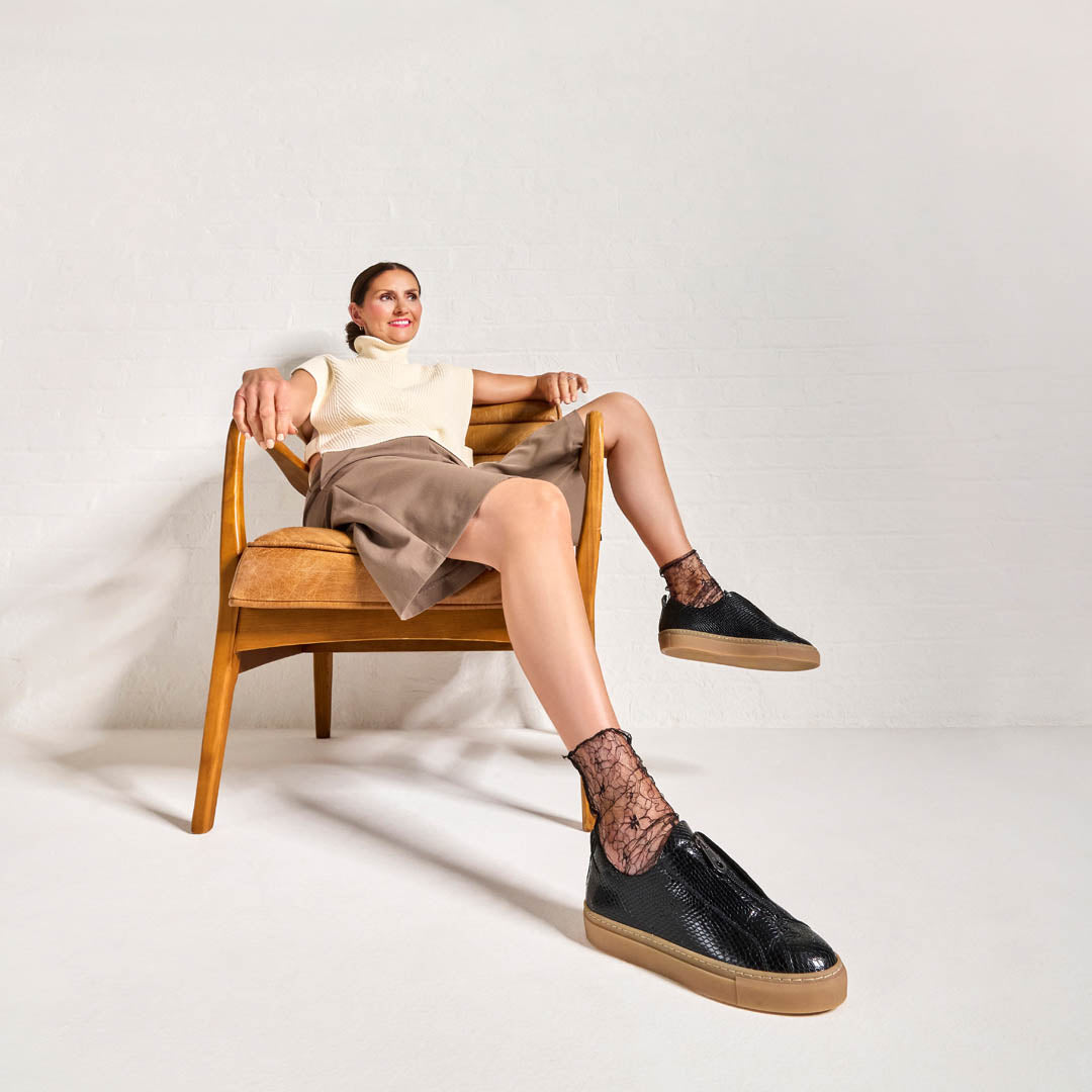 Woman sitting on a chair wearing black Estella sneakers with brown soles.