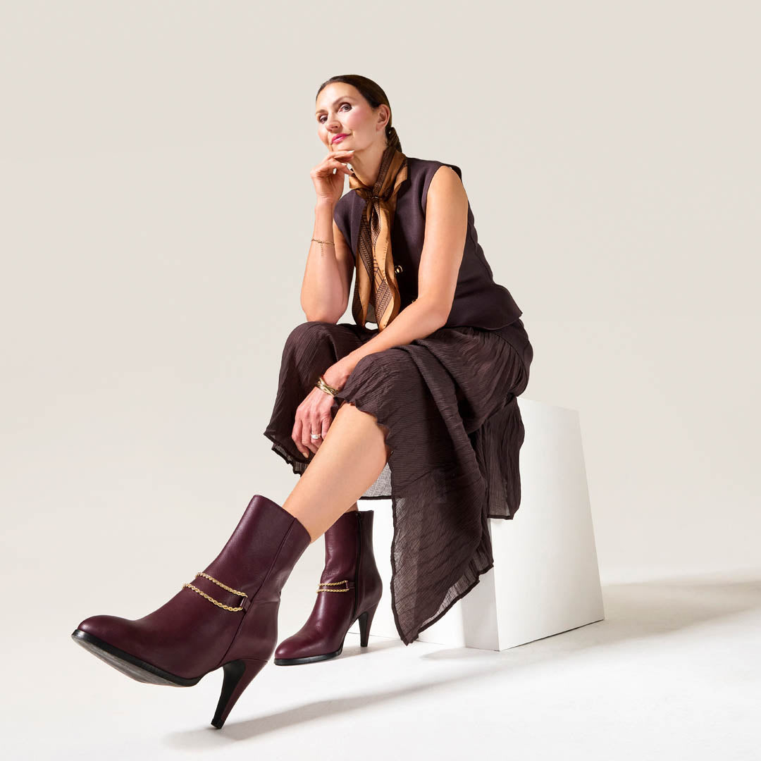 Woman wearing Cherry Maya boots for bunions with gold buckles sitting on a white stool against a light background