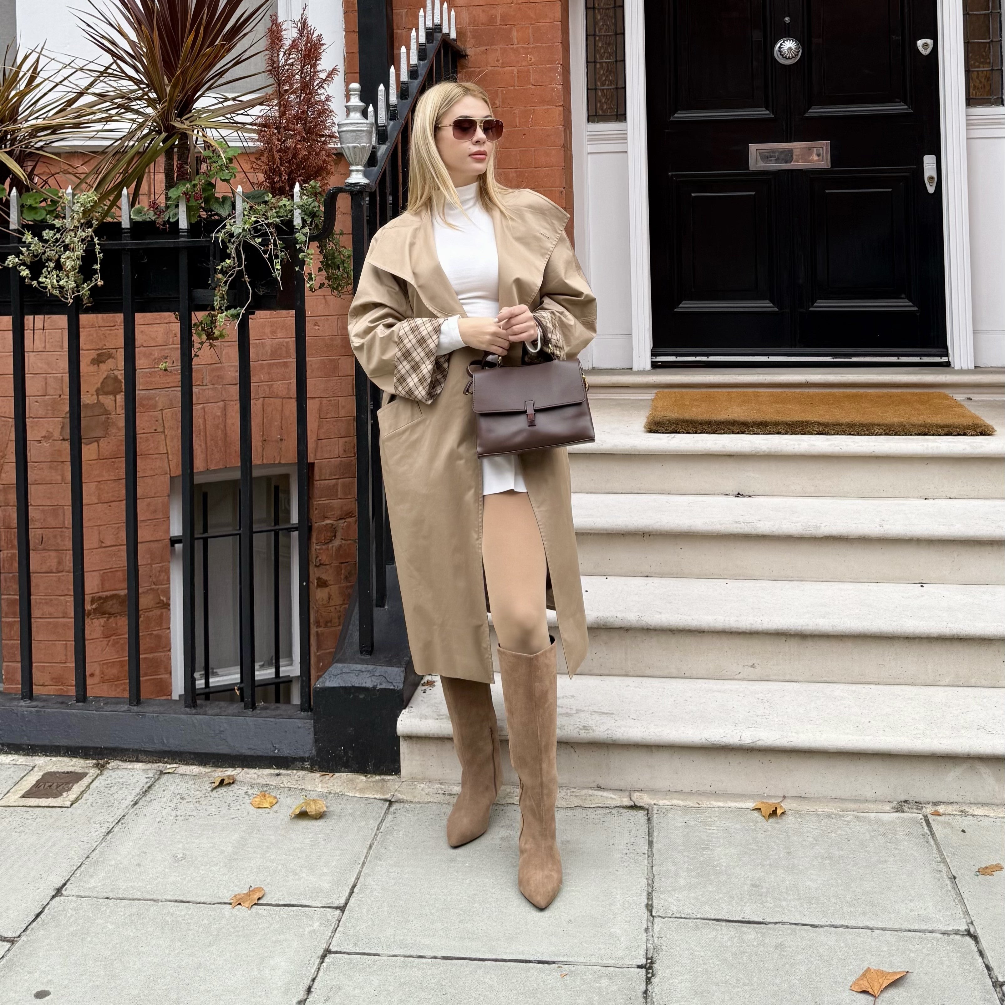 Woman in a beige trench coat and knee-high boots standing on a sidewalk.