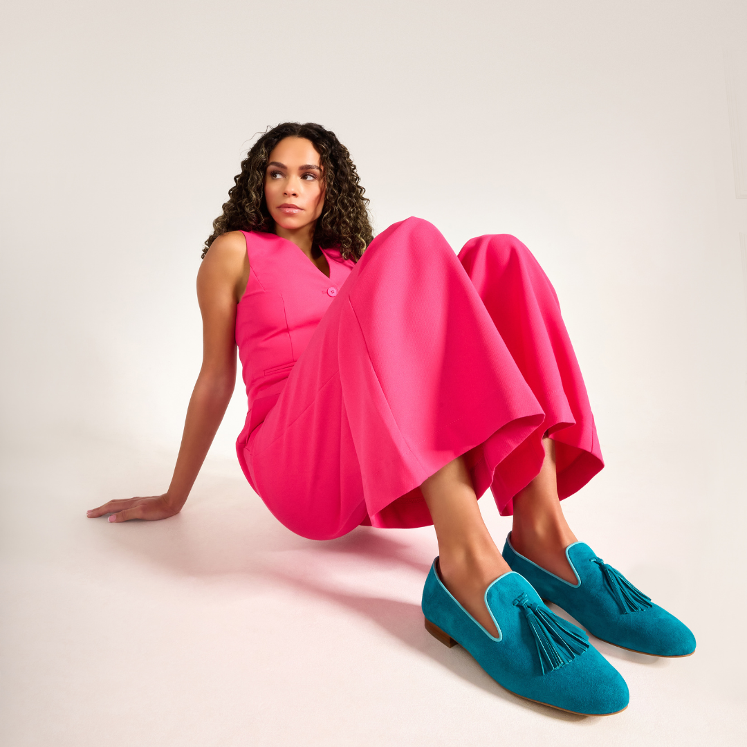Woman wearing a bright pink dress and blue loafers sitting on a white background