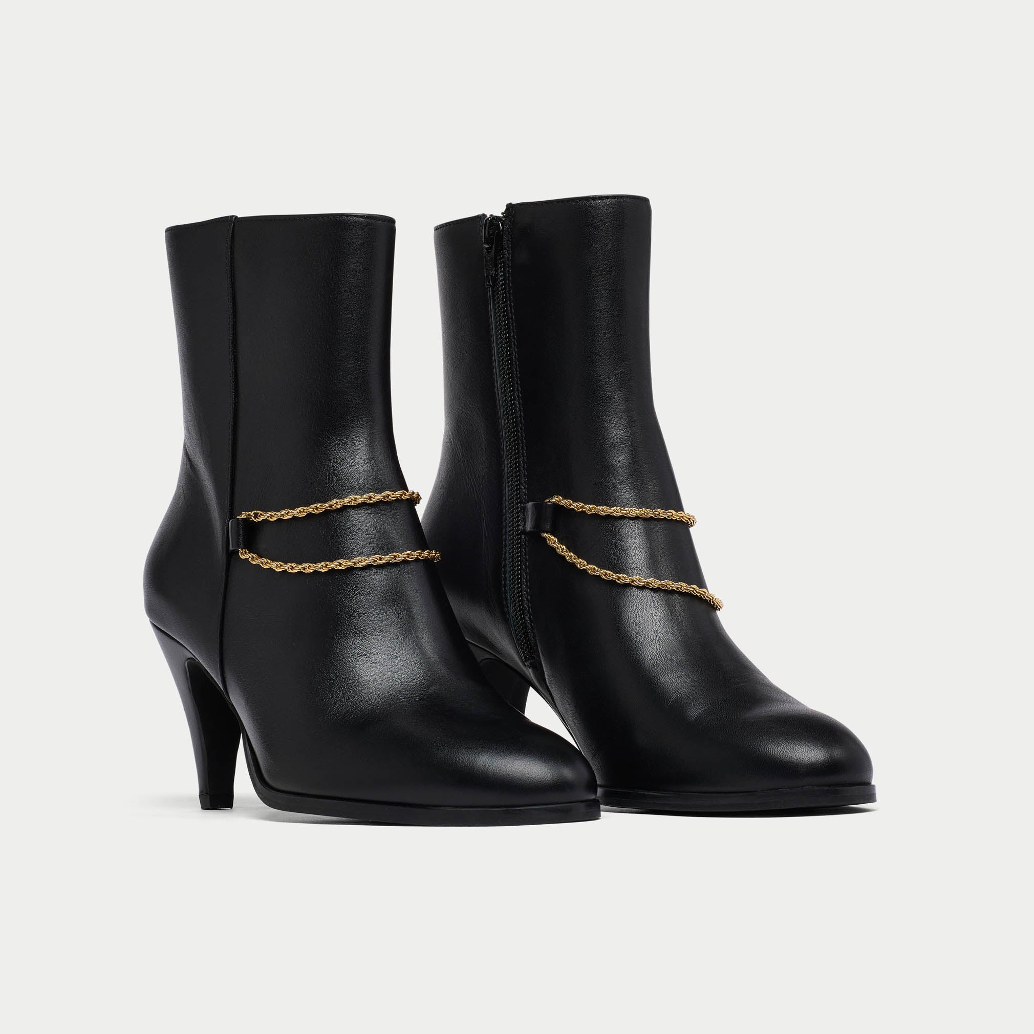 Pair of Maya black ankle boots with gold chain details on a light gray background