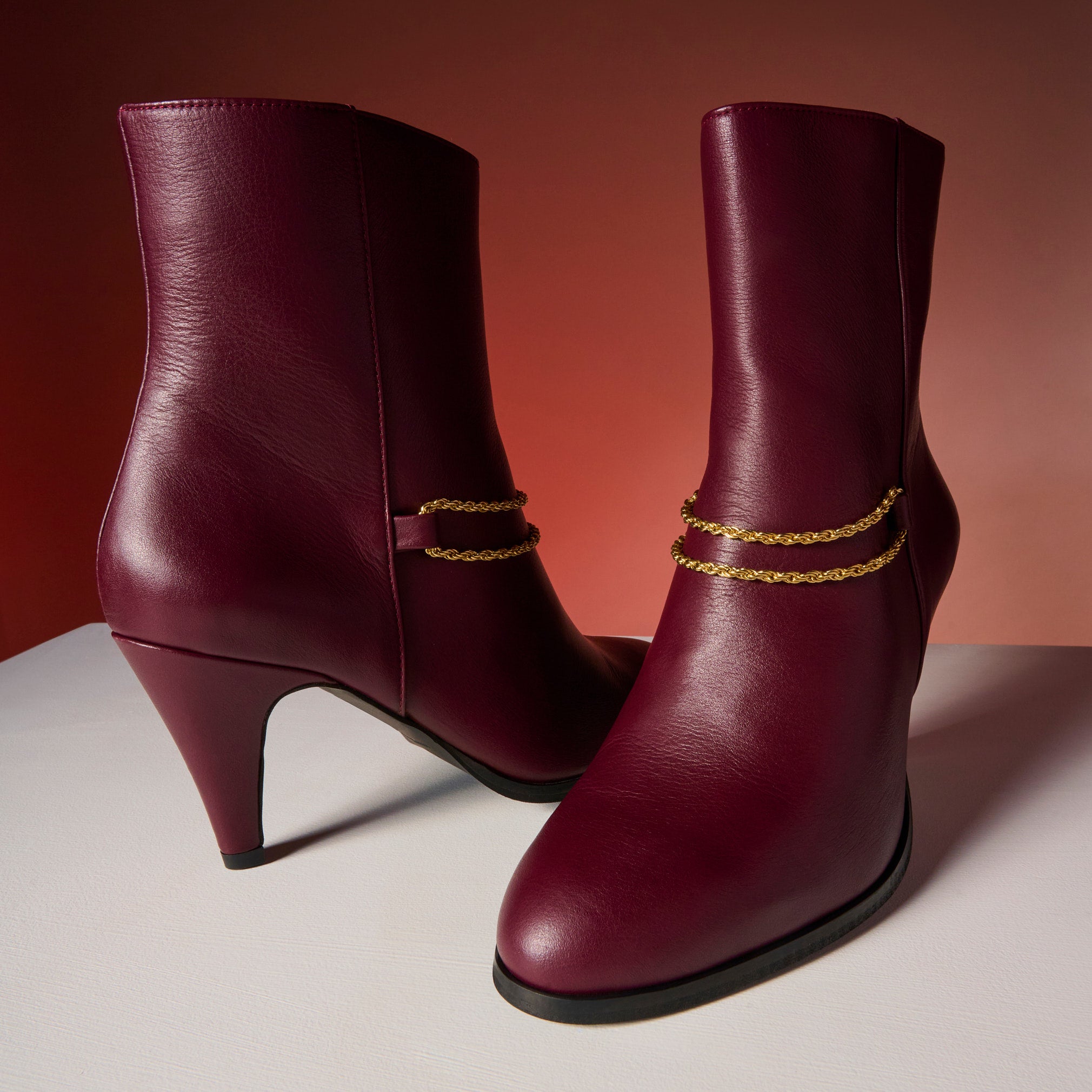 Pair of Maya cherry ankle boots with gold chain details on a gradient background