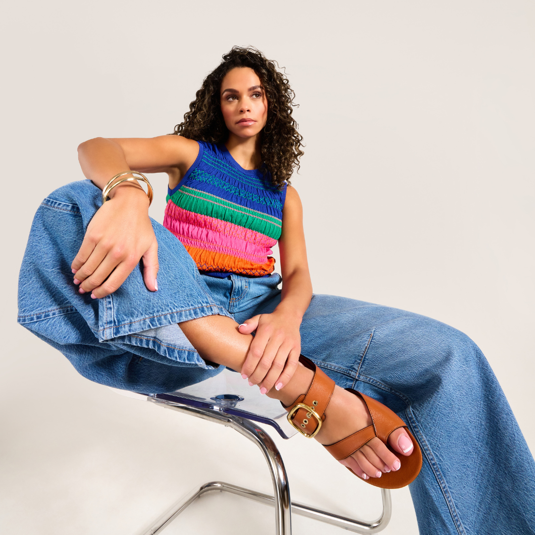 Woman wearing a colorful sleeveless top and blue jeans sitting on a chair.