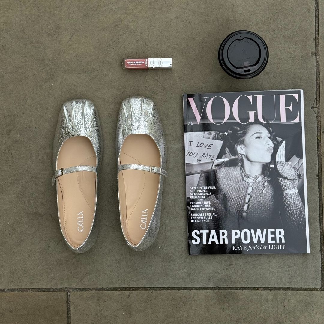 Silver shoes, a magazine titled 'Vogue', a coffee cup, and a lipstick on a textured surface.