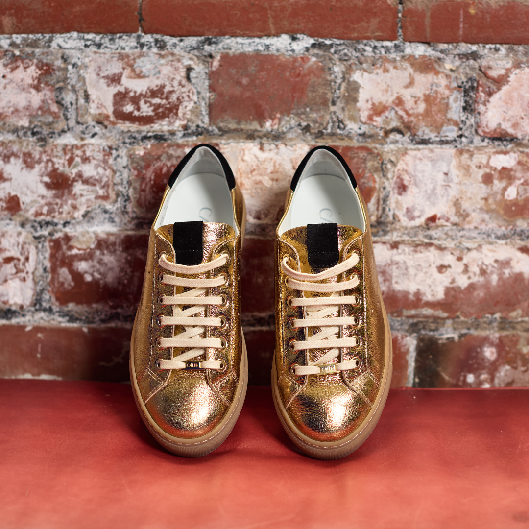 Pair of gold sneakers with white laces on a red surface against a brick wall.
