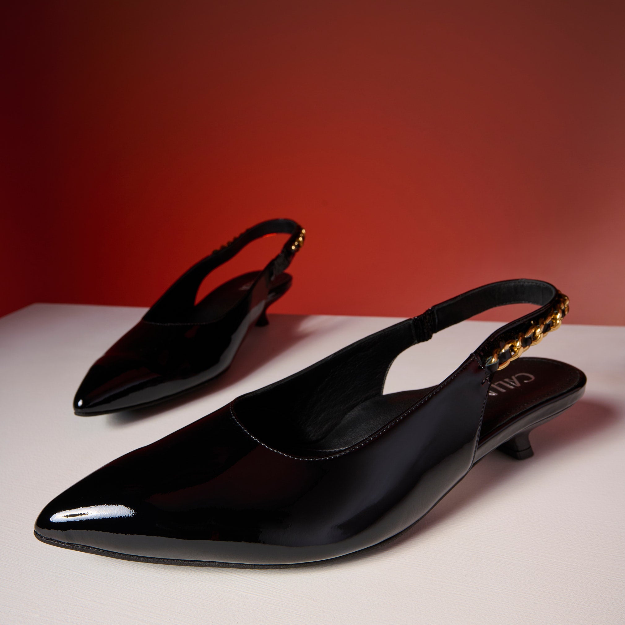 Black patent leather slingback Thea shoes on a white surface with a red background